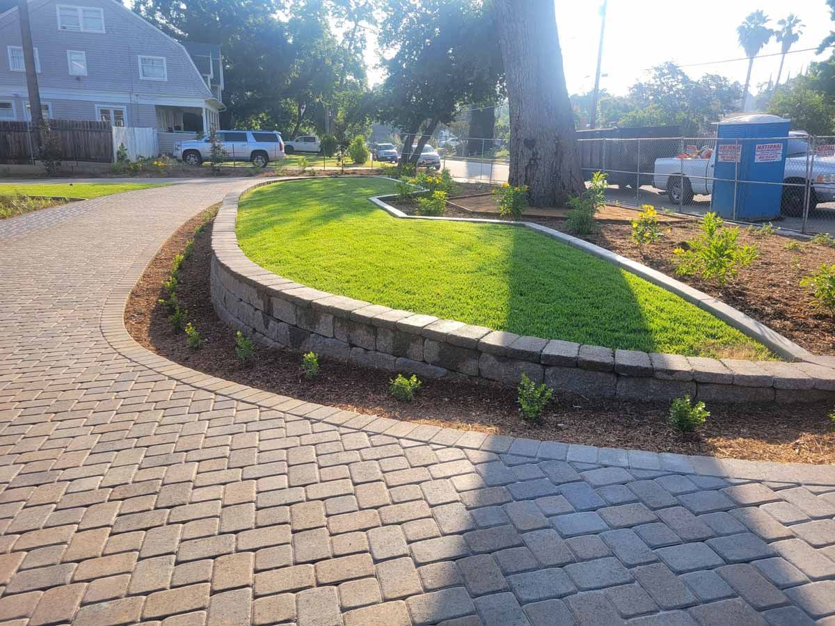 Curving brick driveway with a raised grass bed, bordered by a low stone wall, next to a road.