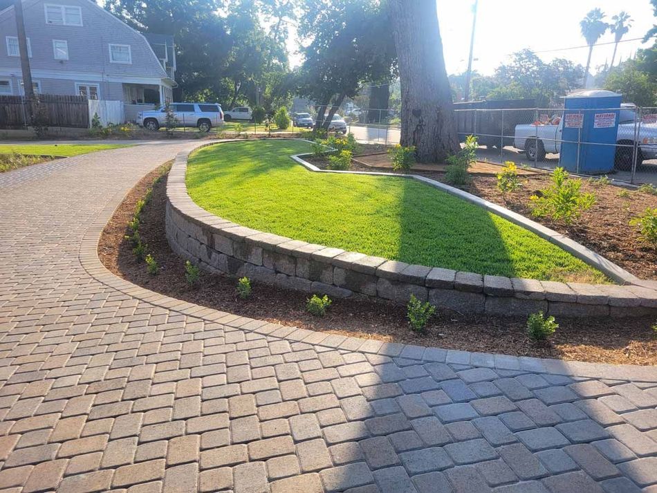 Brick driveway curves around a small lawn and retaining wall with a tree.
