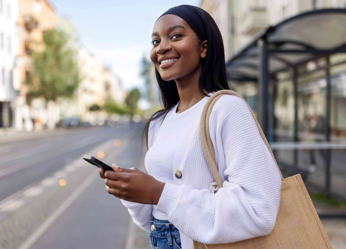 A woman is standing on the sidewalk holding a cell phone and smiling.