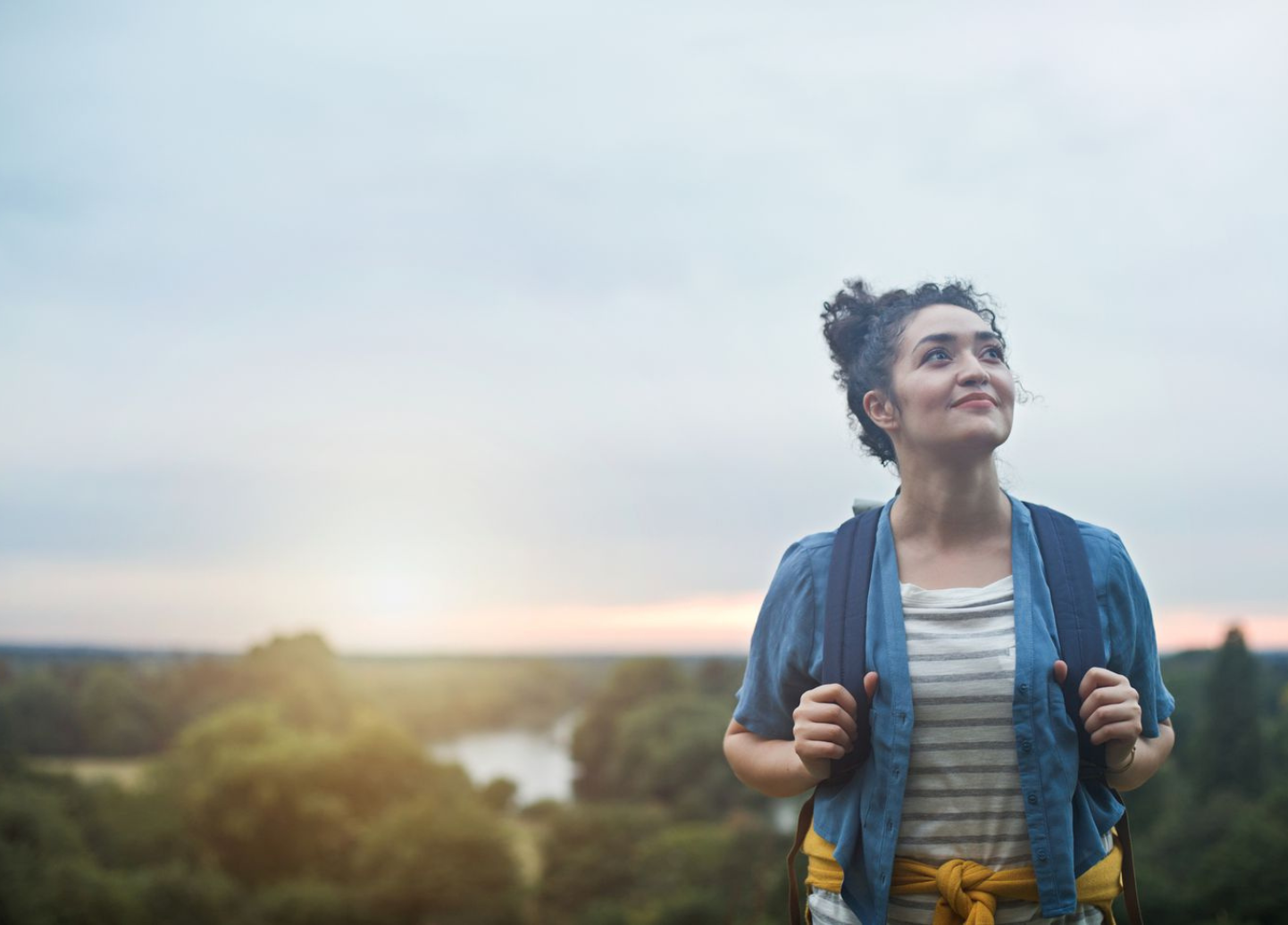 A woman with a backpack is standing on top of a hill looking up at the sky.