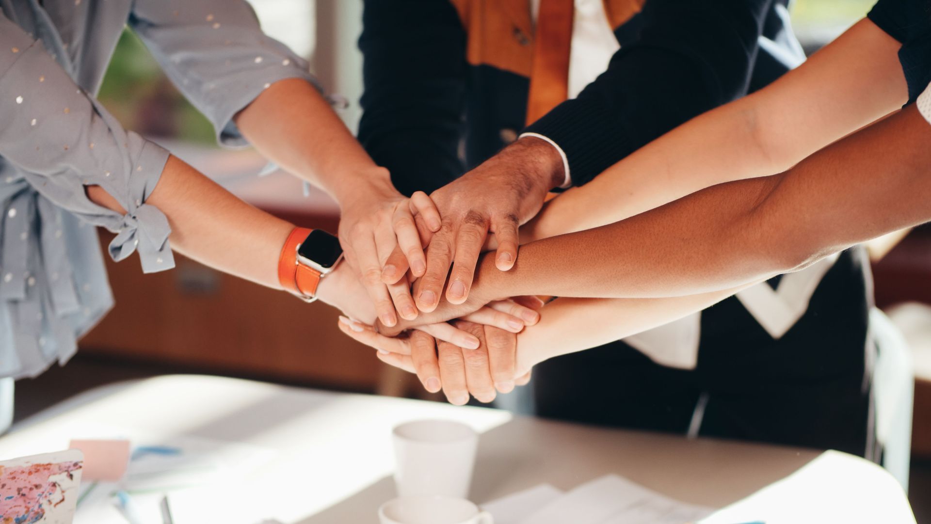 Hands stacked together in a team huddle over a table in a bright office.