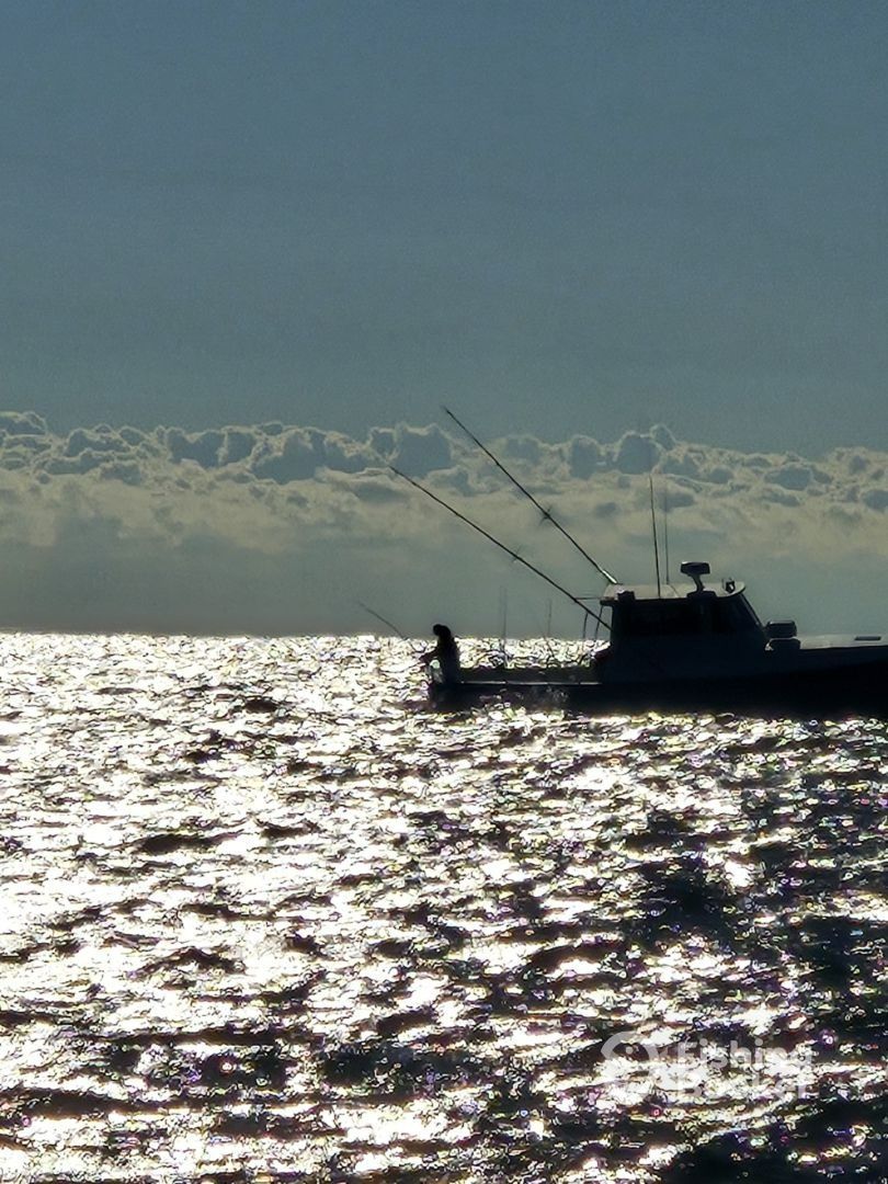 Boat silhouetted on sunlit water with person fishing under a cloudy sky.
