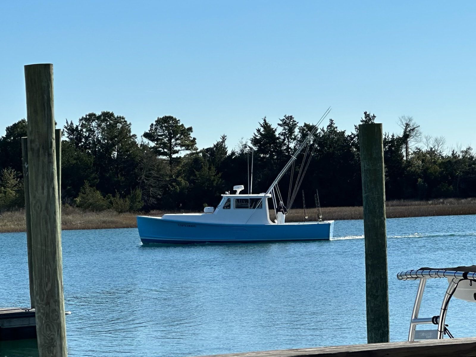Light blue and white boat on water, heading past dock pilings, with trees in background.