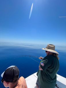 Person fishing from a boat on a sunny day. Blue sky and ocean. Another person is nearby.