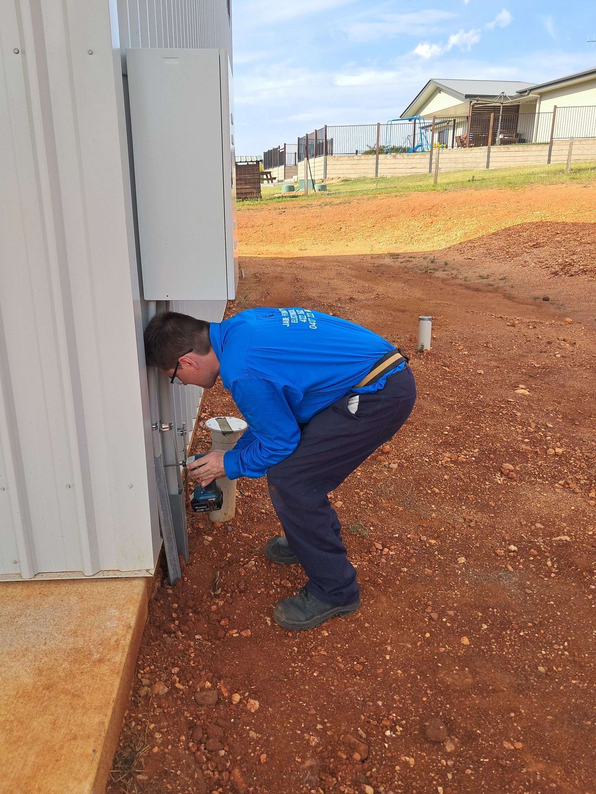 Person in blue workwear installing something on a metal building, outdoors — Jade Fluerty Electrical In Tinana, QLD