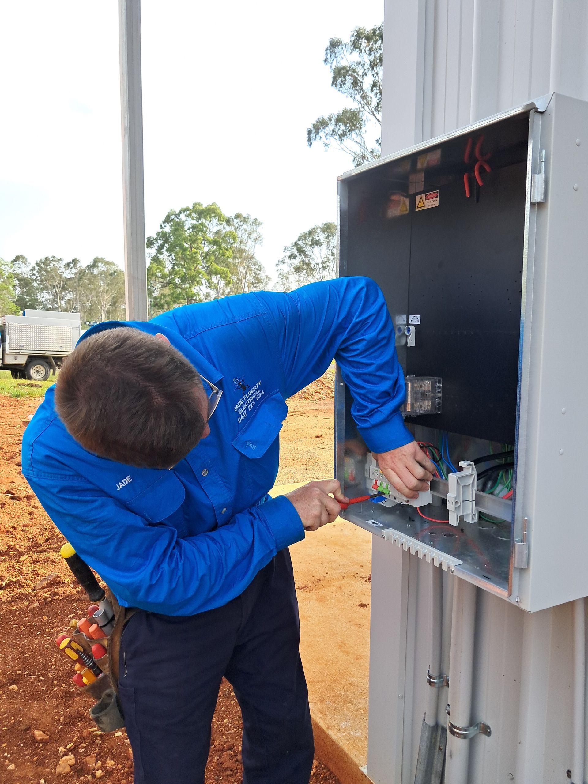 Electrician in blue shirt working on wiring inside a metal electrical box outdoors — Jade Fluerty Electrical In Tinana, QLD