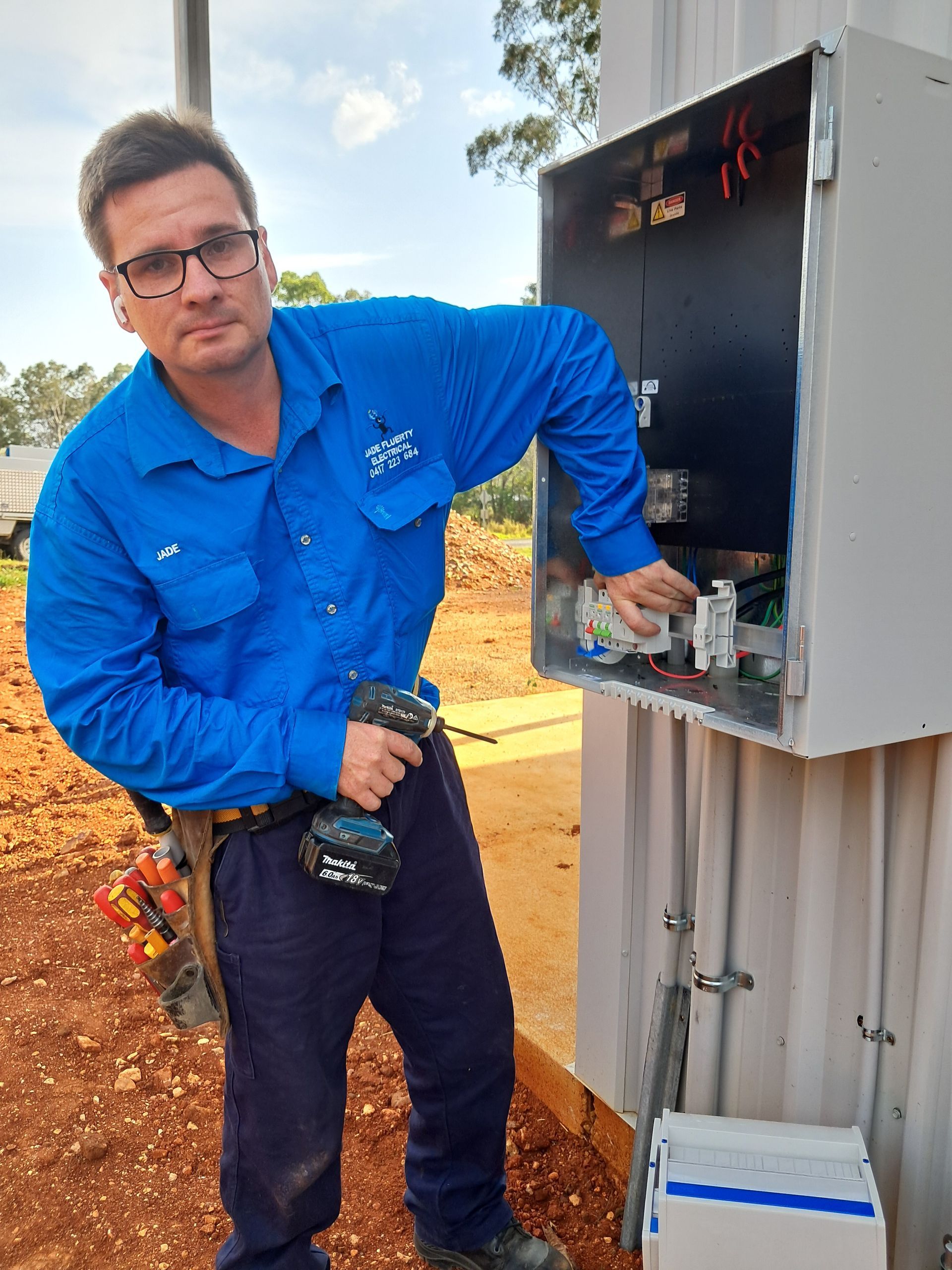Electrician working on a gray electrical box outdoors, holding a drill. He is wearing blue work clothes and glasses — Jade Fluerty Electrical In Tinana, QLD