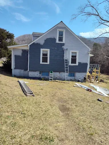 A house under renovation with partially painted dark gray siding, featuring ladders and construction debris in the yard.