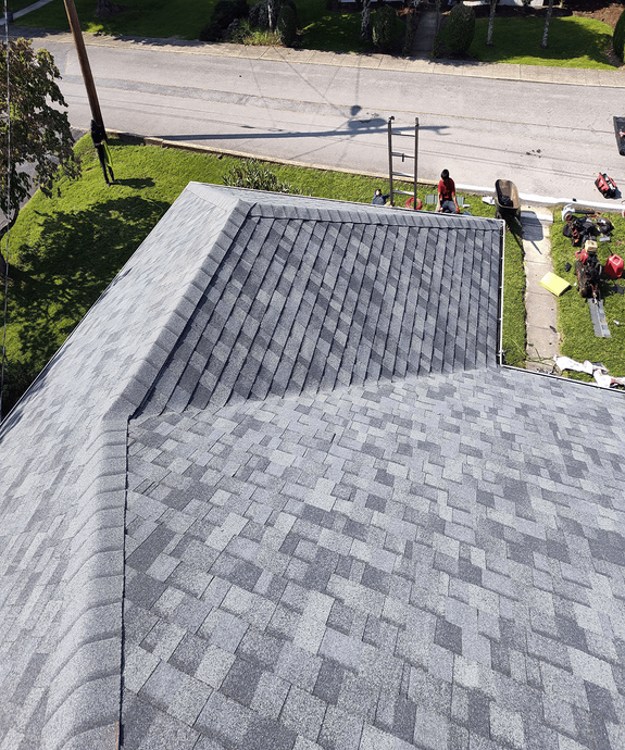 An elevated view of a newly shingled gray roof on a sunny day, with a yard and street visible in the background.
