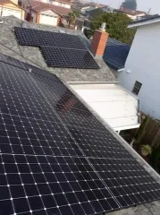 Multiple rows of black solar panels installed on a shingled roof with a brick chimney and a white house nearby.