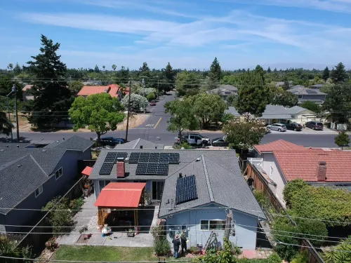 Aerial view of a gray house with solar panels on its roof and a red patio cover, located in a quiet suburban neighborhood.
