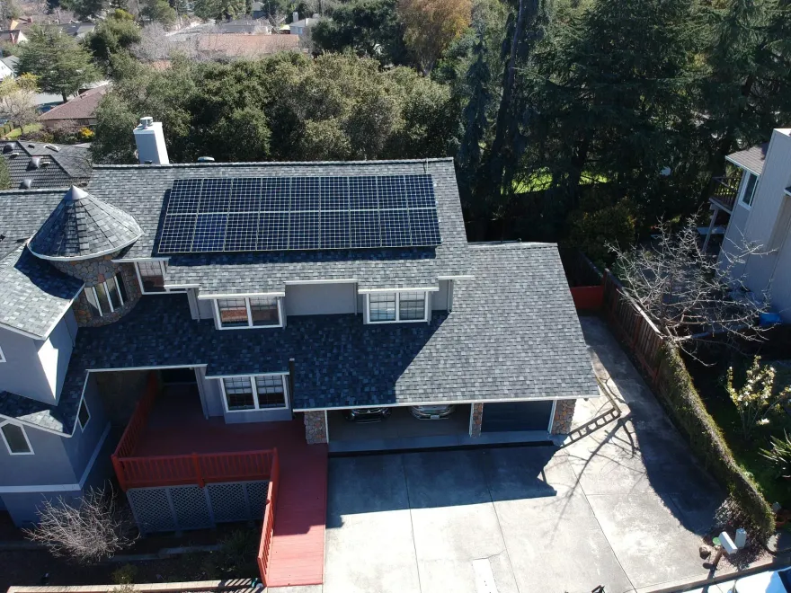An aerial view of a gray residential house with a shingled roof, solar panels, and a red deck.