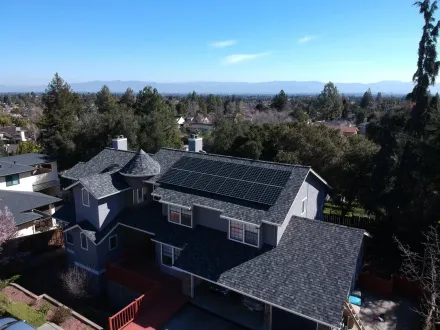An aerial view of a gray, multi-story suburban home with a dark shingled roof and solar panels, set in a sunny residential area.