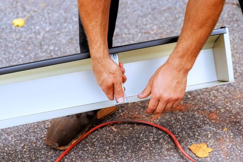 A person works on a metal gutter section, securing a connector piece with a tool while holding it over a paved surface.