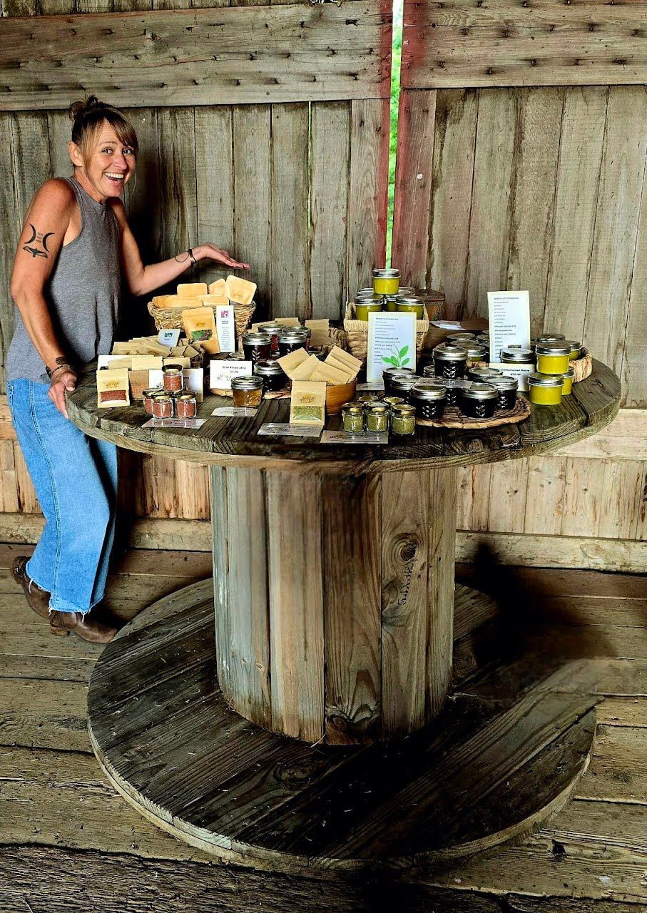 Woman stands next to a wooden cable spool table with food items on display inside a rustic wooden structure.