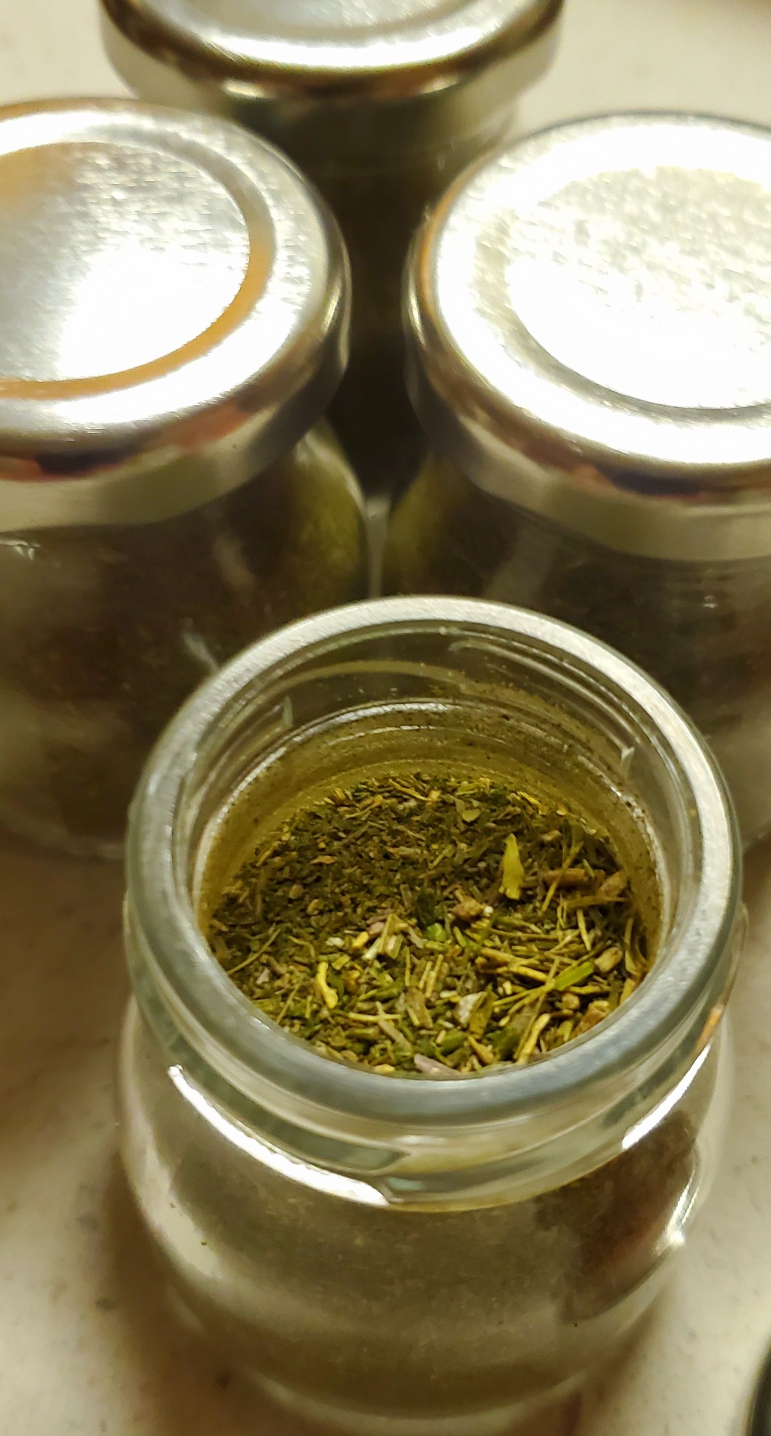 Small glass jars filled with dried herbs, sitting on a light surface. One jar is open.