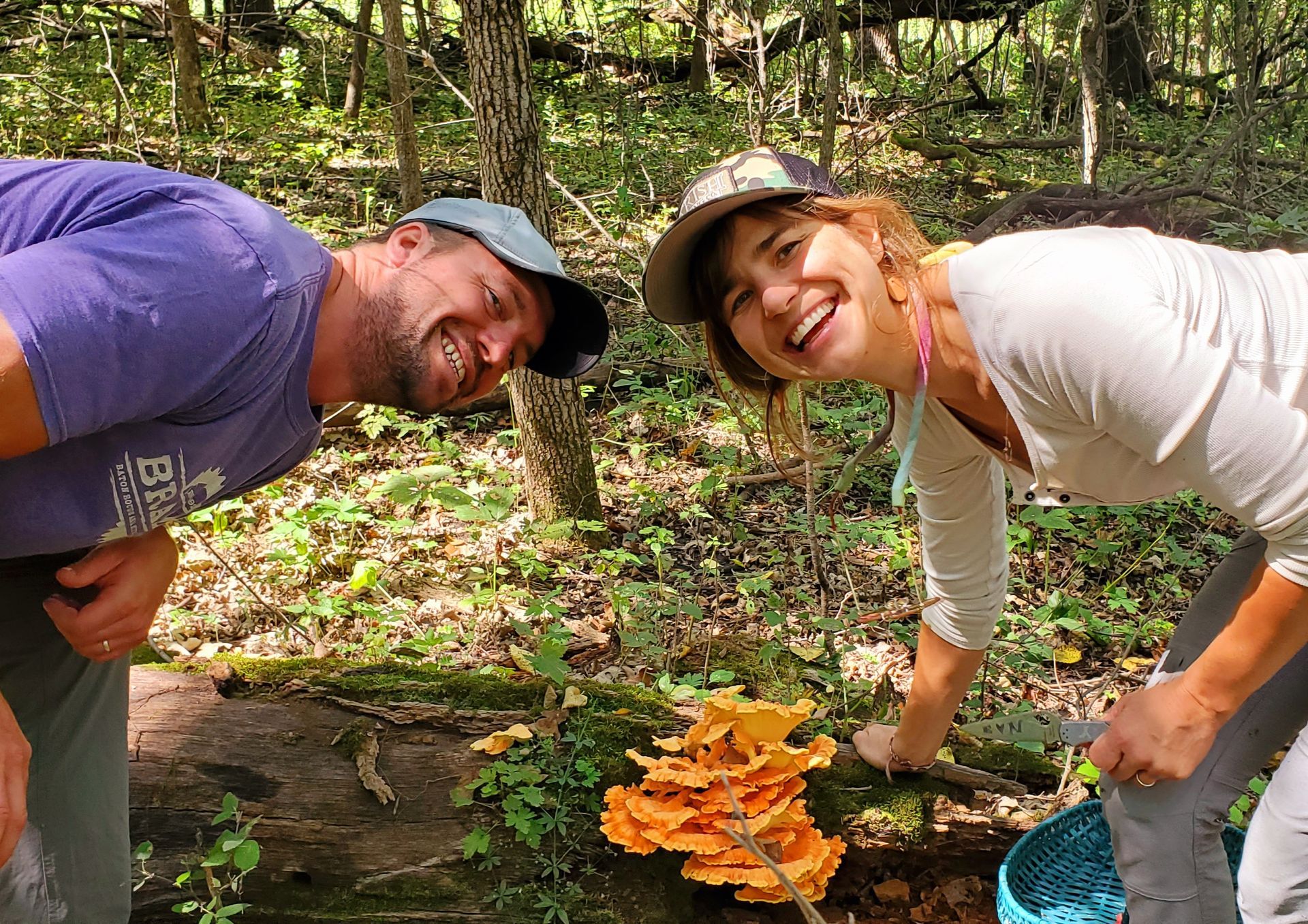 Two people smile, inspecting orange mushrooms on a log in a forest.