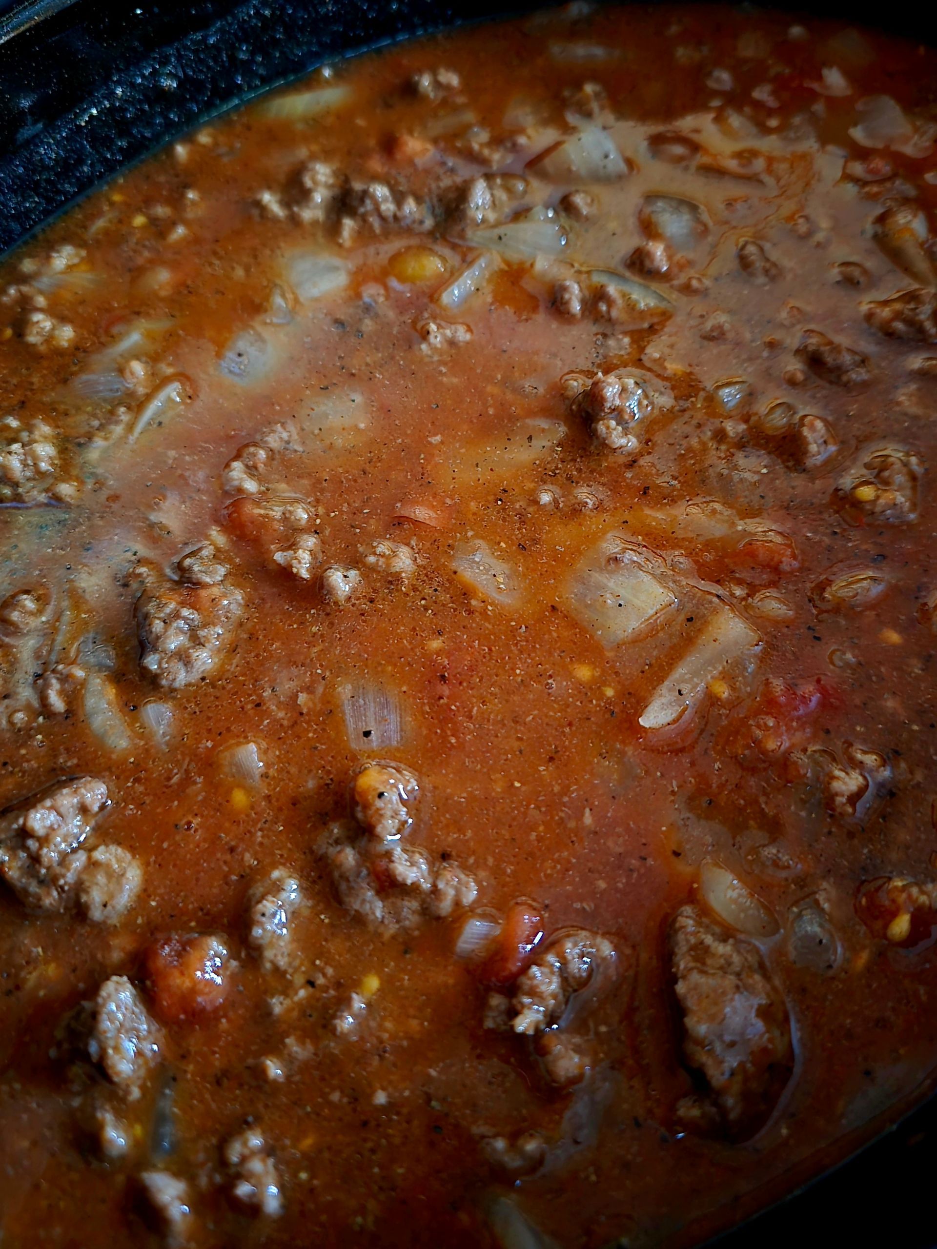 Beef and vegetable stew simmering in a dark pot. Red-brown sauce with visible meat chunks and onions.
