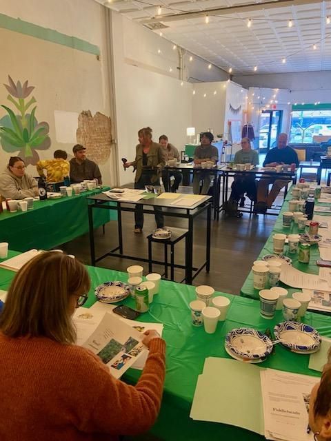 People at a table listening to a speaker. Meeting in a bright room with green tablecloths and paper cups.