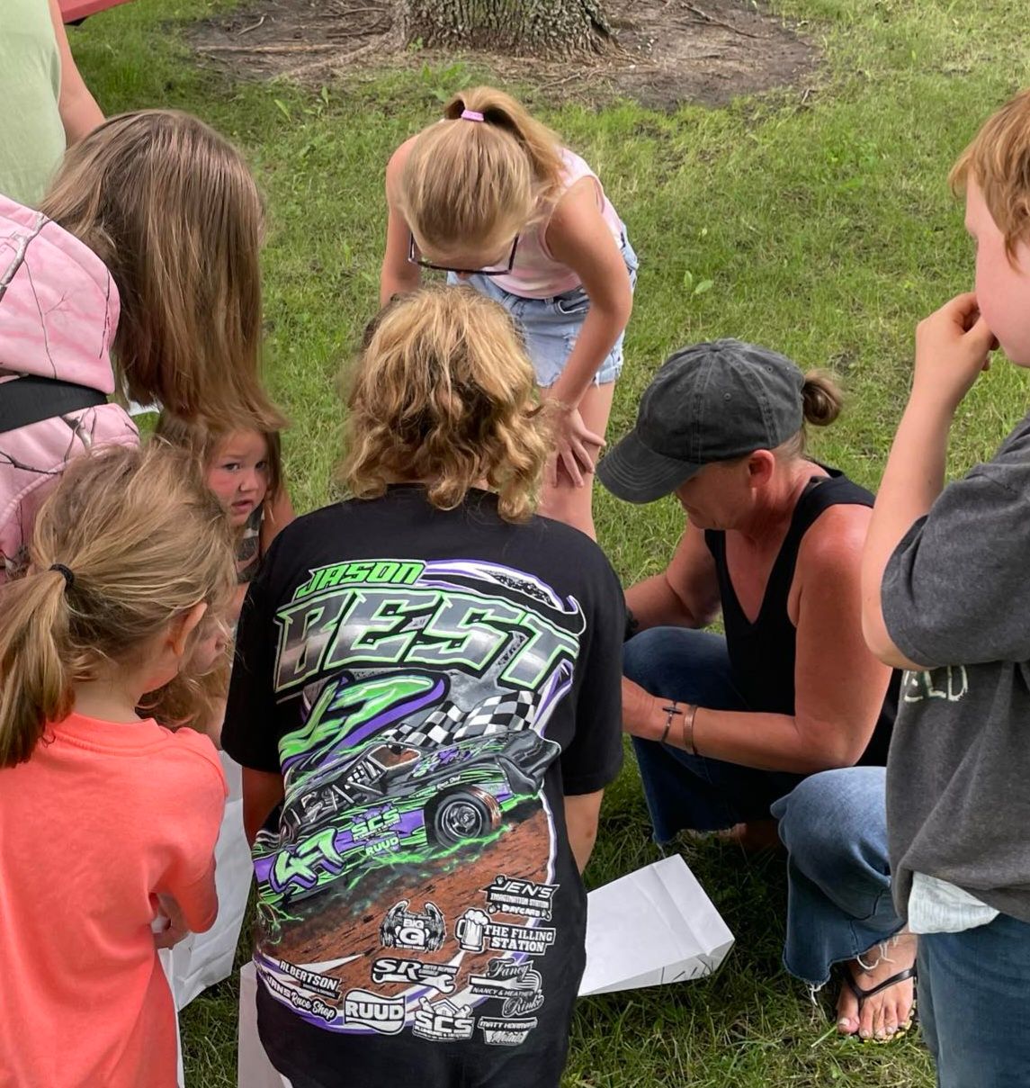 People gathered outside, looking at something on the ground. A woman in a race car shirt is in the center.