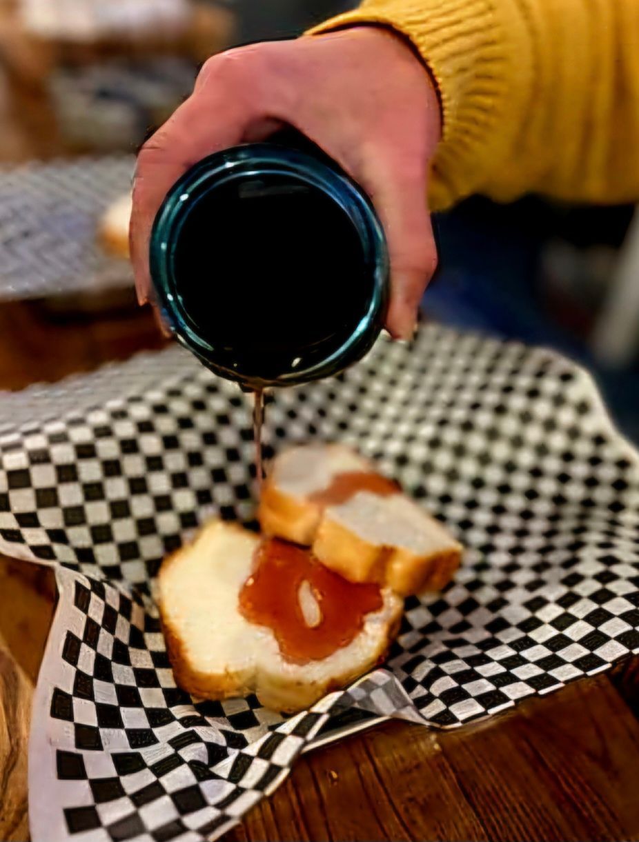 Person pouring dark syrup from a blue container onto bread slices with jam on a checkered paper-lined basket.