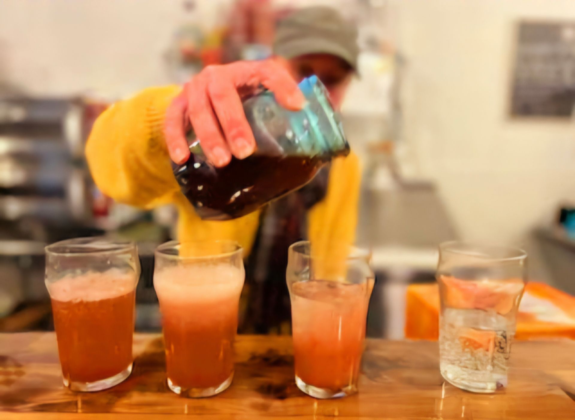 Bartender pouring dark liquid into three glasses with pink liquid, fourth glass of water, wooden bar.