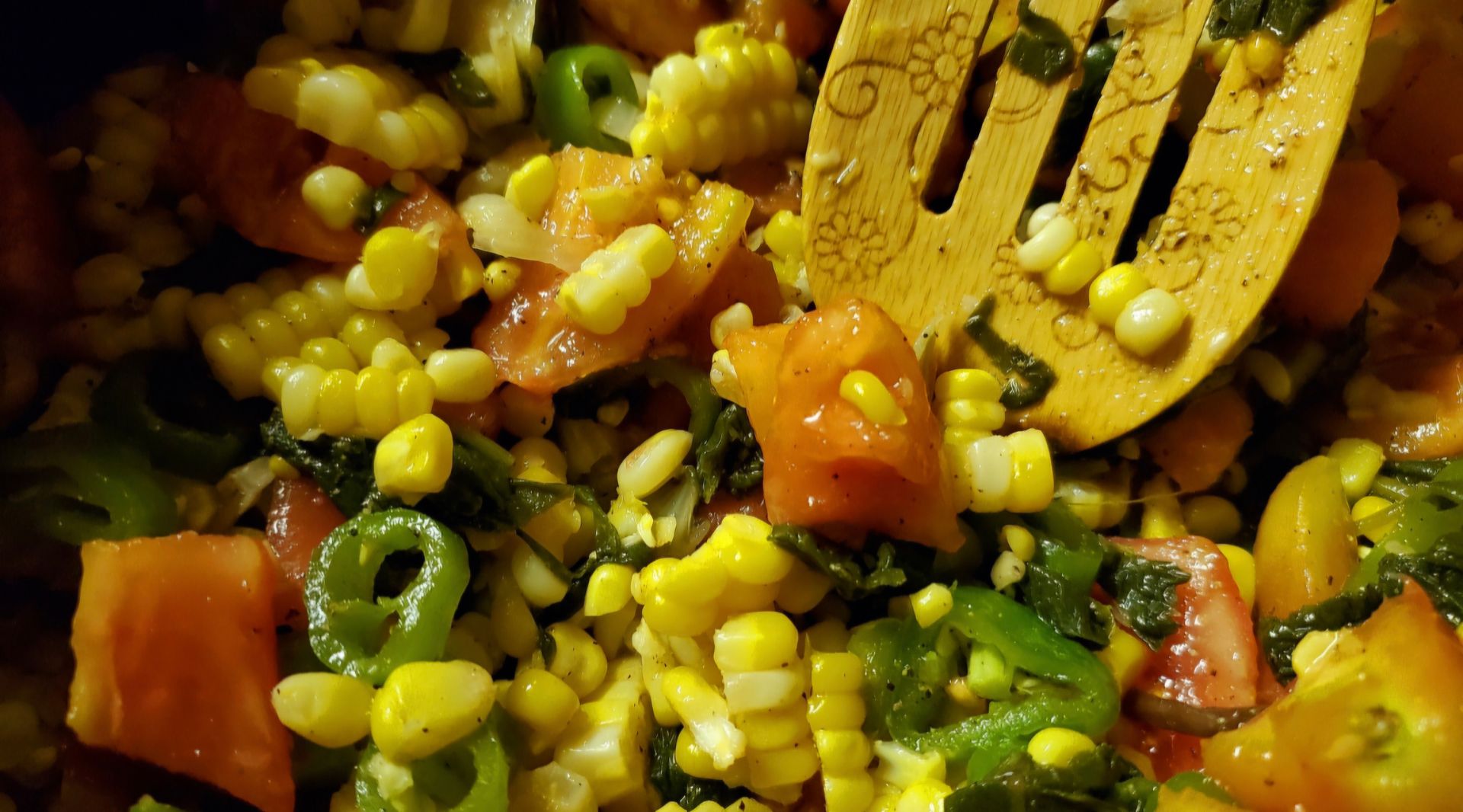 Corn and tomato salad with green peppers, shown in a bowl with wooden serving spoon.