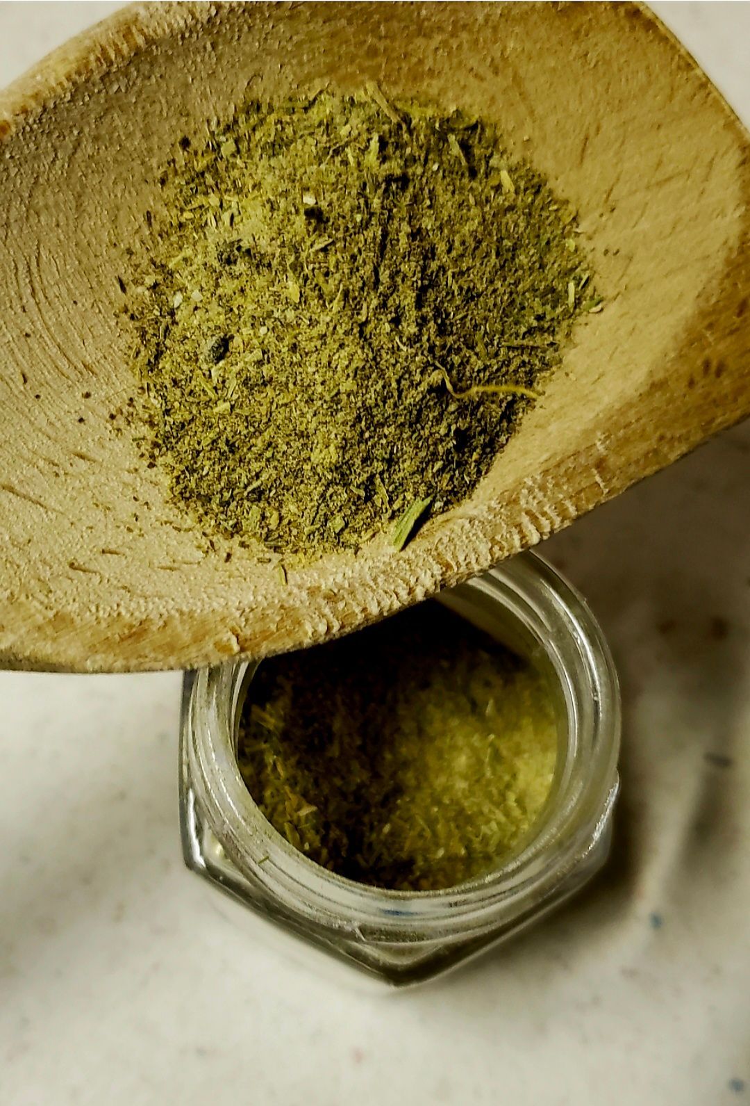 Green powder being poured from a wooden spoon into a small glass jar.