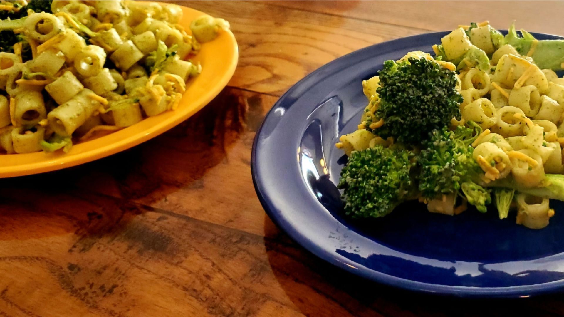 Two plates of pasta with broccoli on a wooden table. One plate is yellow, the other blue.