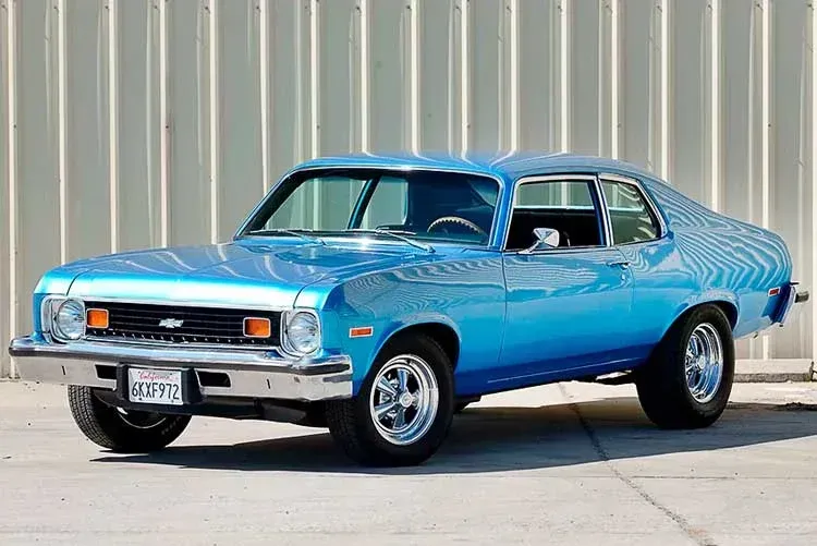 A bright blue 1970s Chevrolet Nova parked on concrete against a ribbed metal wall.