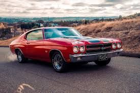 A red 1970 Chevrolet Chevelle SS parked on an asphalt road in a hilly landscape under a cloudy sky.