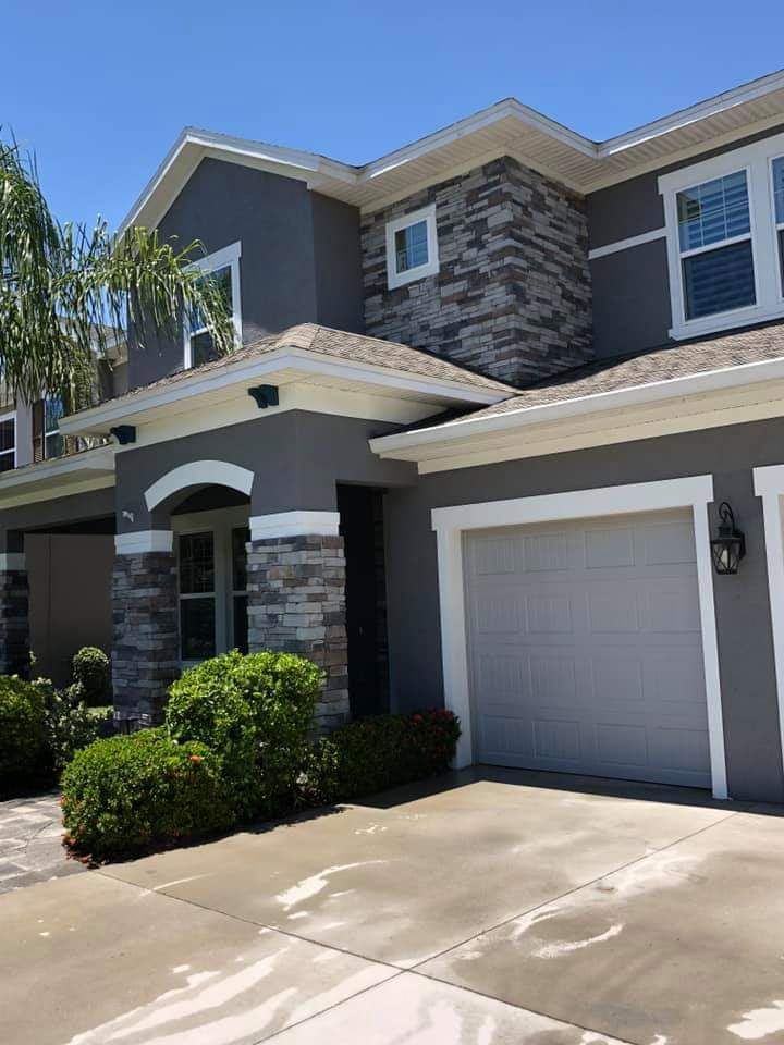 A two-story grey stucco house with stone accents, white trim, a one-car garage, and green landscaping under a clear sky.