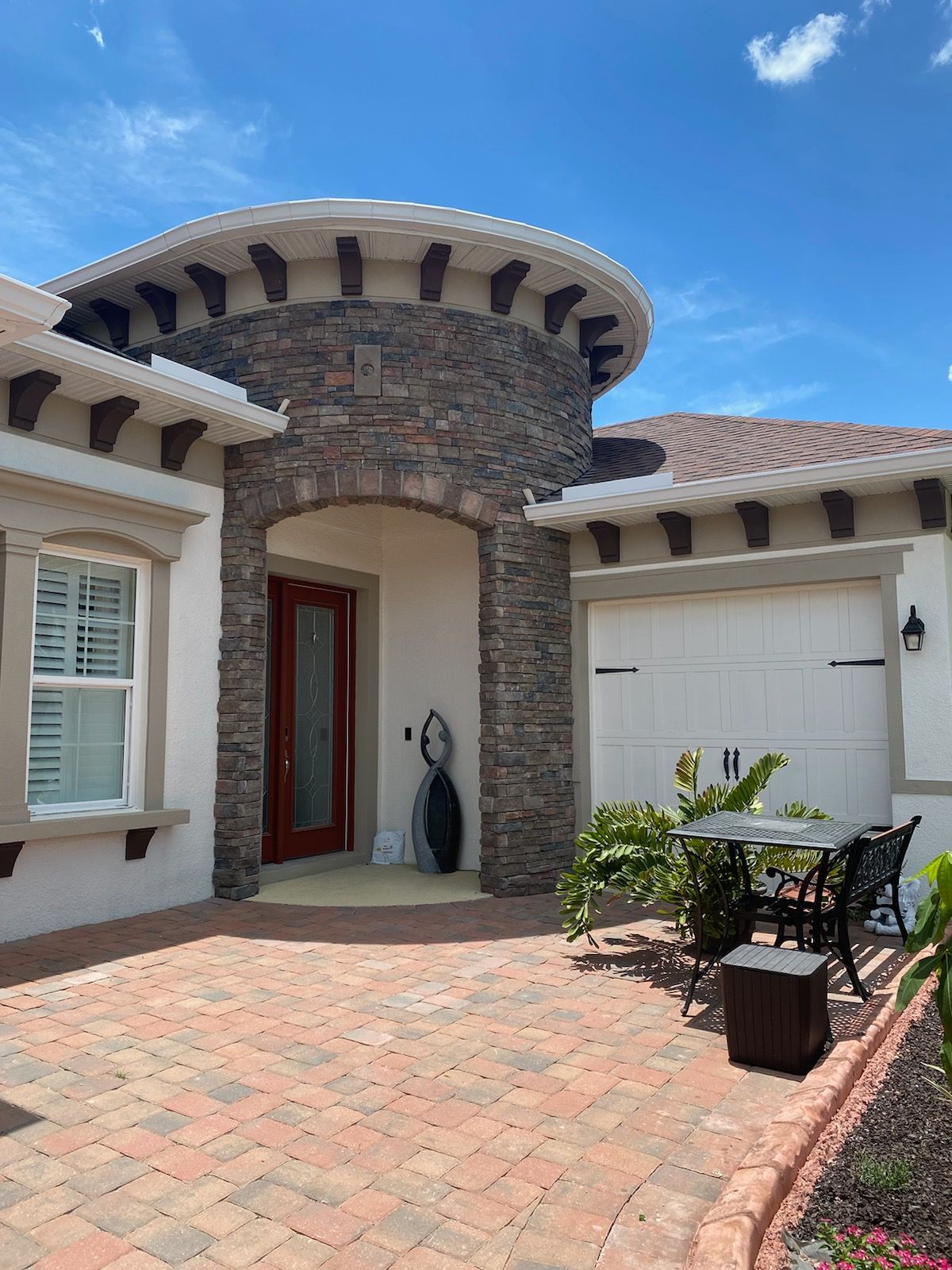 A house exterior featuring a stone-faced arched entry, a tan garage, and a brick paver patio with bistro furniture.