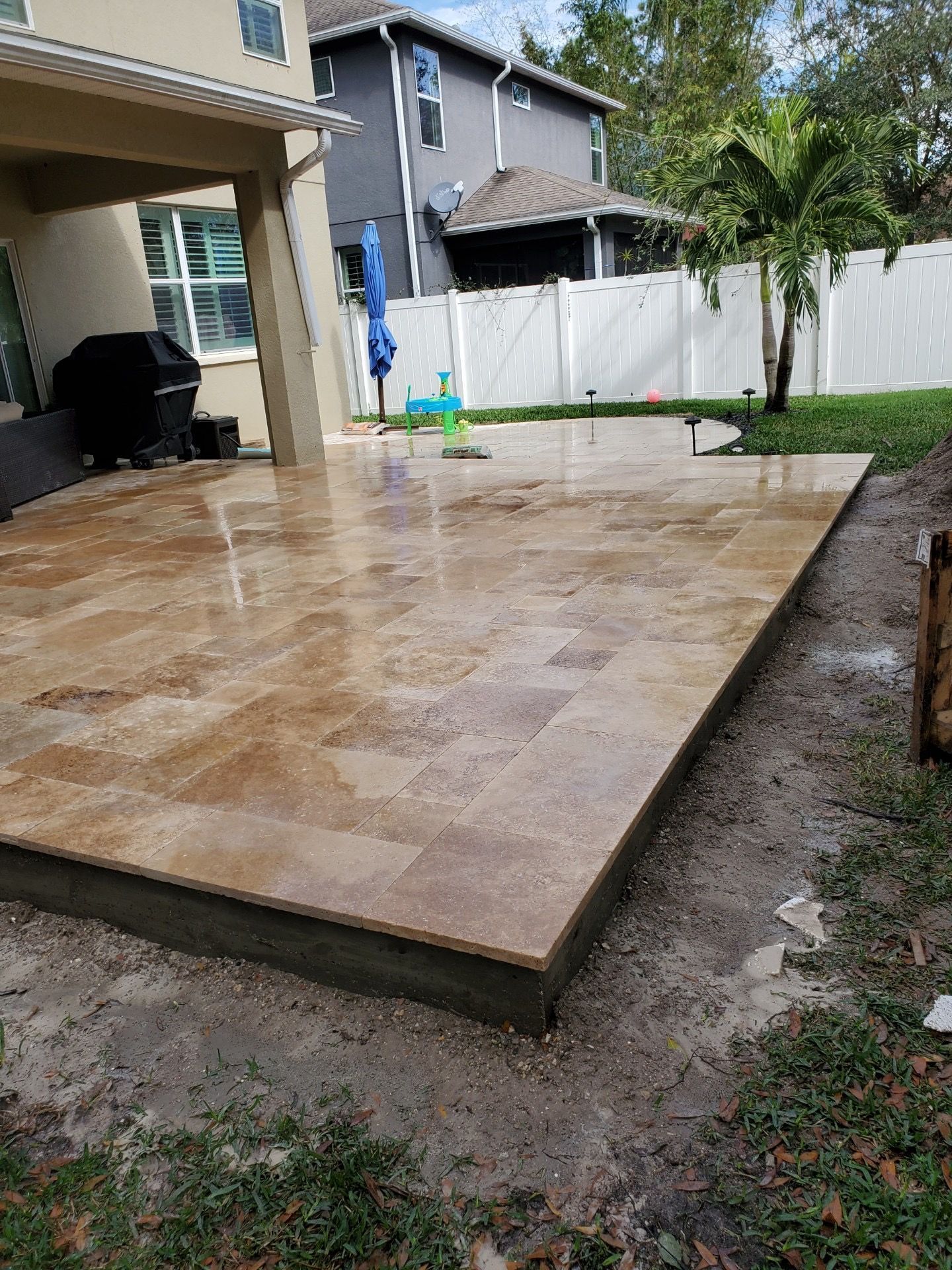 A newly installed tan travertine patio extension on a residential backyard lawn beside a multi-story house.
