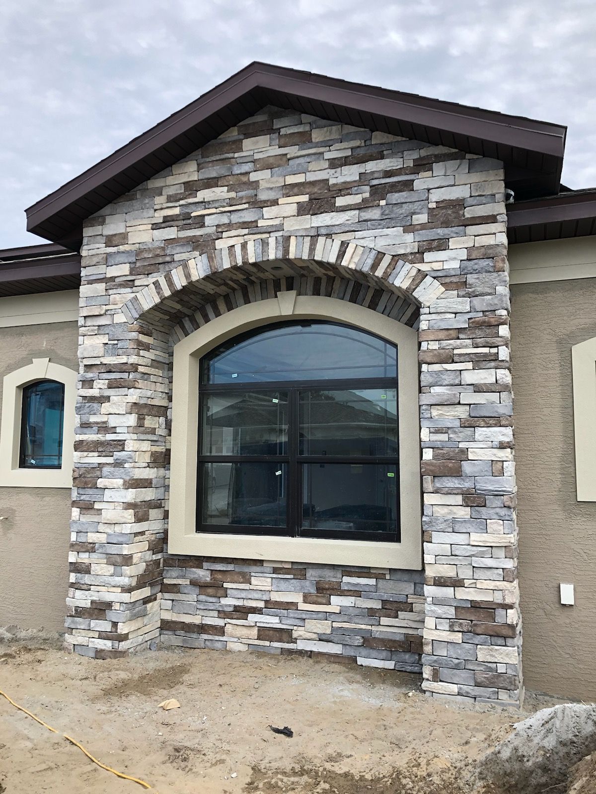 A stone-veneered exterior house wall featuring an arched window with a cream-colored frame and a dark roof gable.