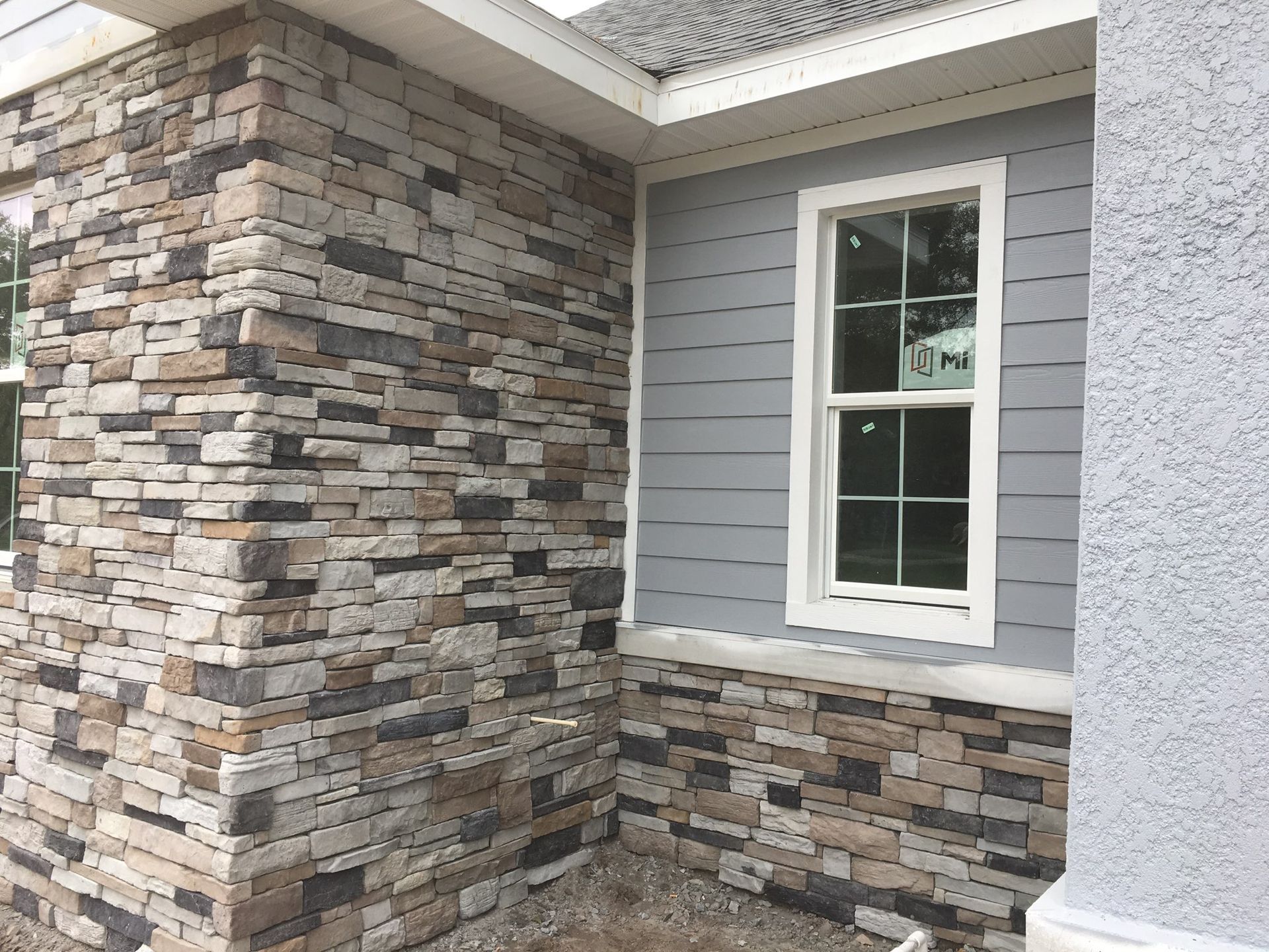 A building exterior with a gray-toned stone veneer corner, gray horizontal siding, a white-framed window, and white stucco.