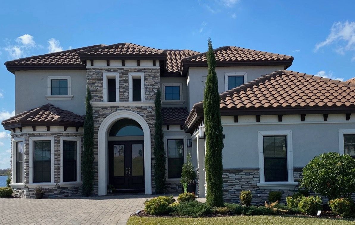 A two-story Mediterranean-style luxury house with a stone facade, arched entryway, and red tile roof under a blue sky.