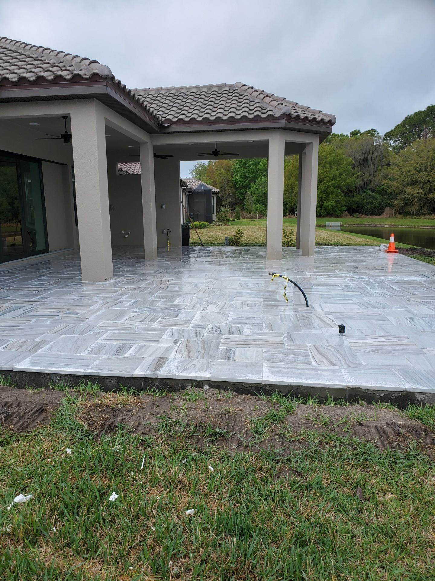 Newly installed light-colored stone patio under a covered lanai attached to a home overlooking a grassy area and pond.