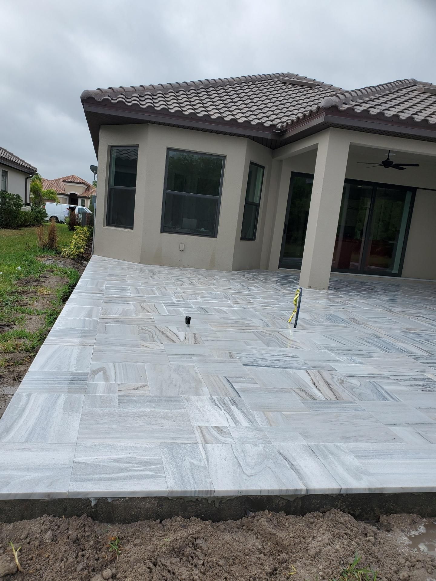 Newly installed gray and white marble paver patio attached to the back of a beige house under a cloudy sky.