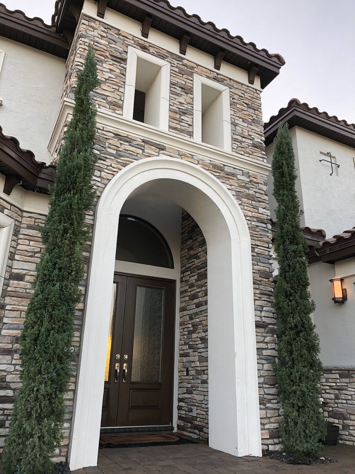 A front entrance of a home with stacked stone siding, an arched white doorway, and two tall, slender evergreen trees.