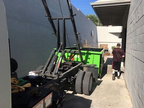 Green dumpster being lifted by a truck. A person walks next to the wall. Bright daylight.