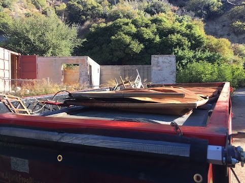 Red flatbed trailer with debris in front of a partially demolished building in a wooded area.