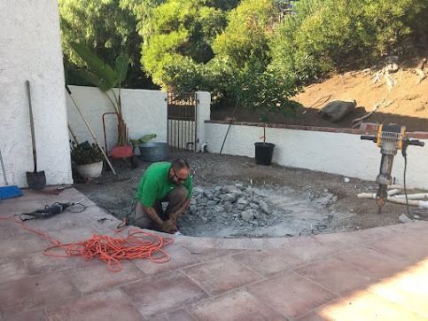 Man breaking up concrete in a patio area surrounded by white walls and greenery.