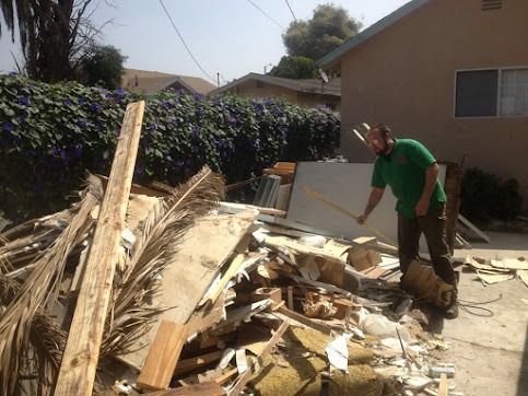 Man in green shirt, brown pants standing near pile of debris, using a tool. Outdoors, sunny.
