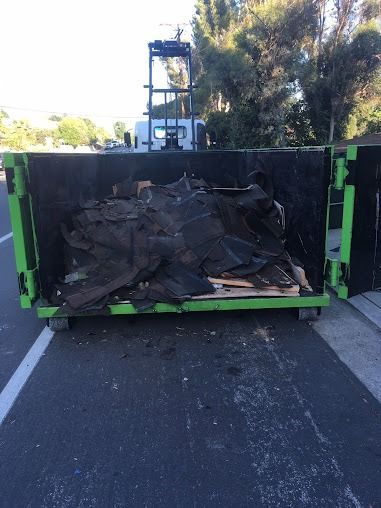 Green dumpster filled with black debris, likely roofing material, on a road. A truck is visible behind it.