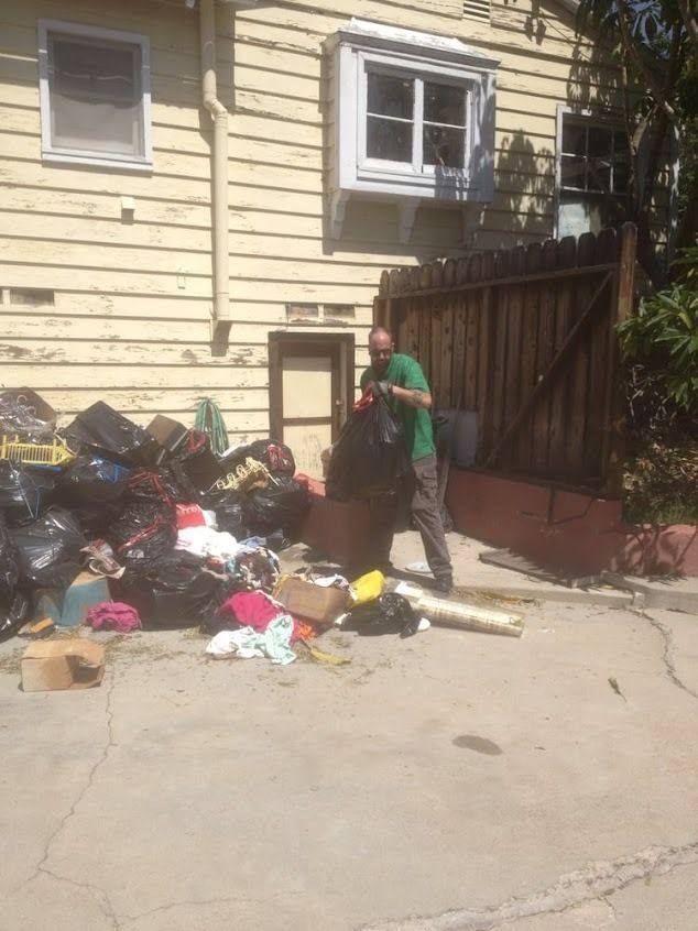 Person loading black trash bags on a concrete area outside a yellow building and a wooden fence.