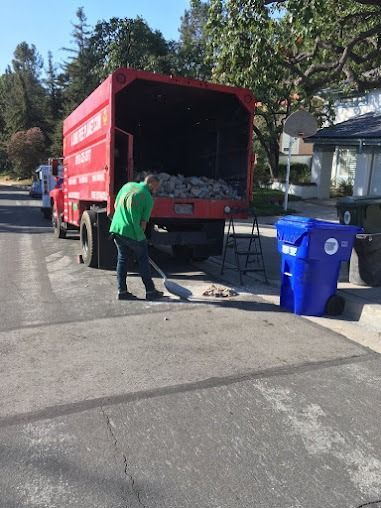 A garbage truck with its back open; a worker sweeps trash from the street. A blue recycling bin is on the right.