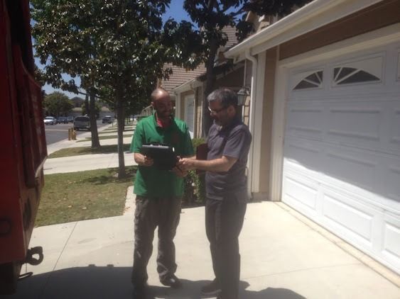 Two men standing on a driveway, one in green shirt holding clipboard, the other in gray shirt, signing it.