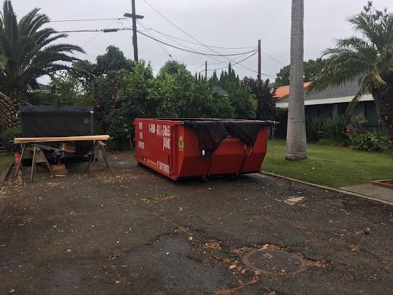 Red dumpster in driveway, covered with black tarp. A sawhorse with a black object sits nearby. Overcast sky.