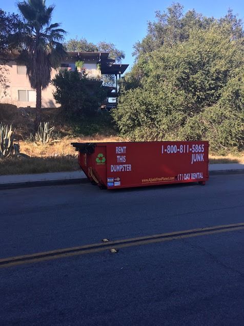 Red dumpster on a street with a phone number for rental, with a residential building and trees in the background.